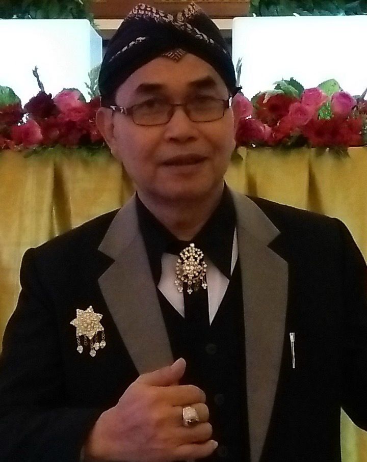 Whatsapp image 20260608 at 21 02 51 catrawarta Man wearing a black suit with gray lapels and a jeweled brooch seated in front of pink flowers